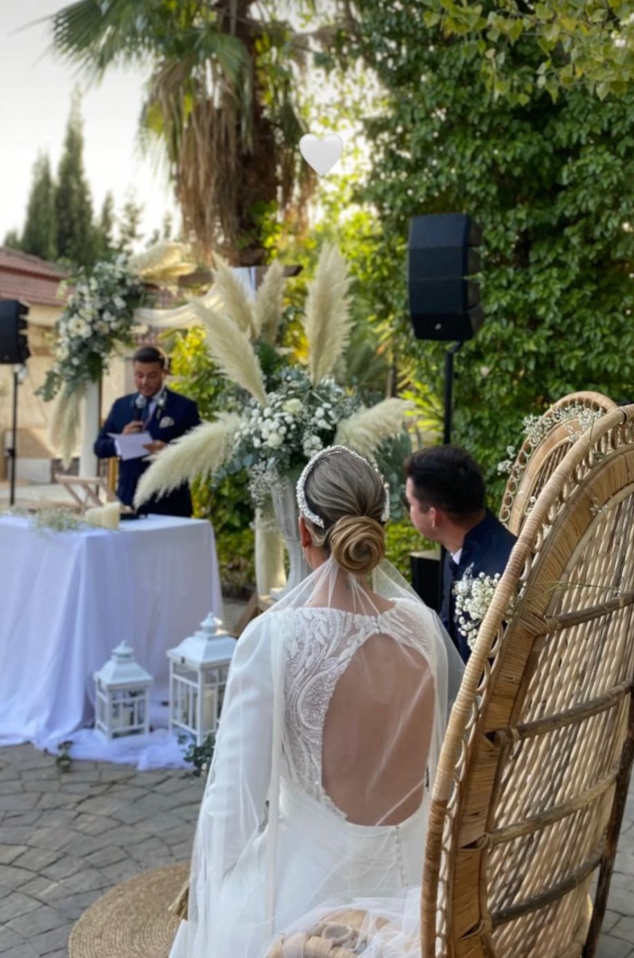 Pareja sentada durante una ceremonia al aire libre, con oficiante al micrófono y un altavoz junto al espacio de celebración